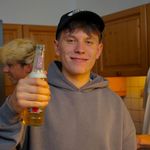 Junger Mann in grauem Hoodie und Baseballcap hält eine Bierflasche und lächelt in die Kamera – gesellige Atmosphäre in einer gemütlichen Küche