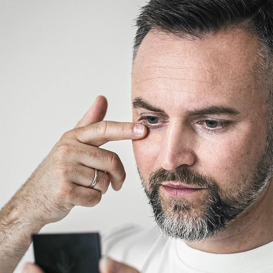 Mann mit gepflegtem Bart trägt Concealer oder Augencreme unter den Augen auf, während er sich in einem kleinen Spiegel betrachtet – Hautpflege und Beauty-Routine für Männer.